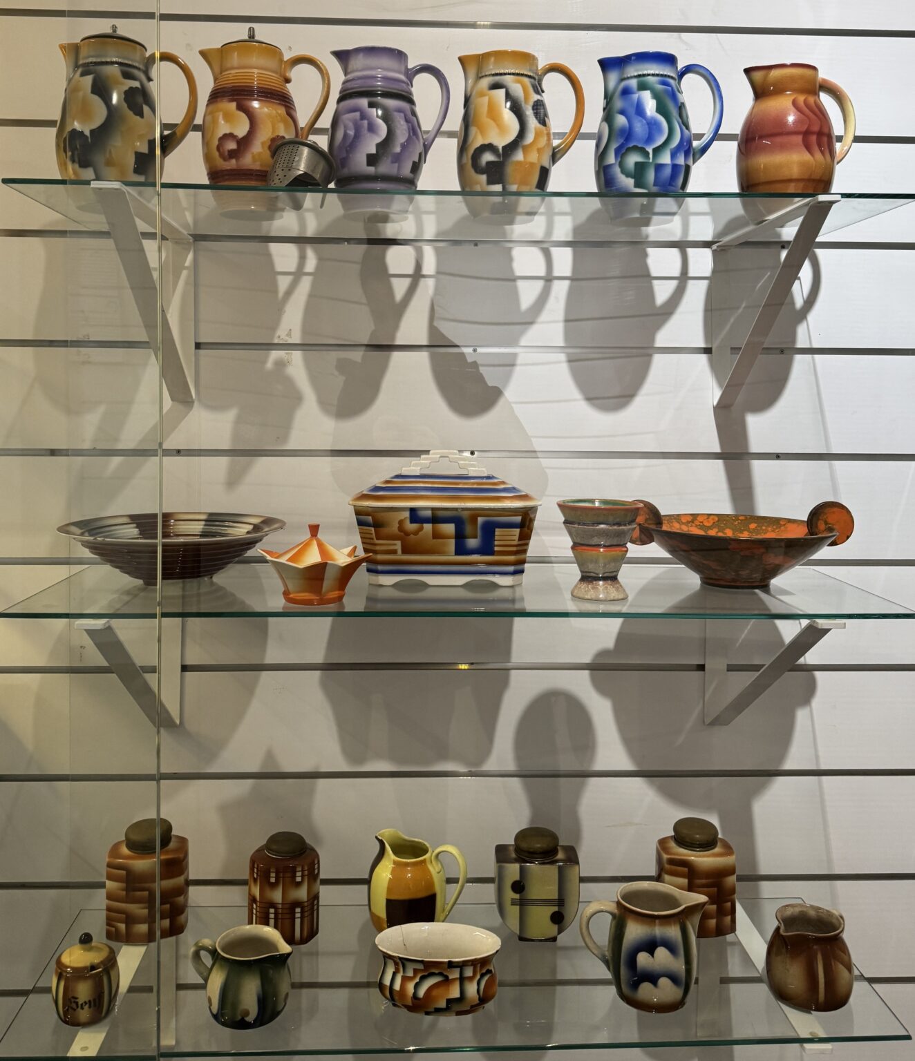 Trzy szklane półki jedna nad drugą, na nich ceramiczne obiekty: kolorowe dzbanki, pojemniki, miseczki.