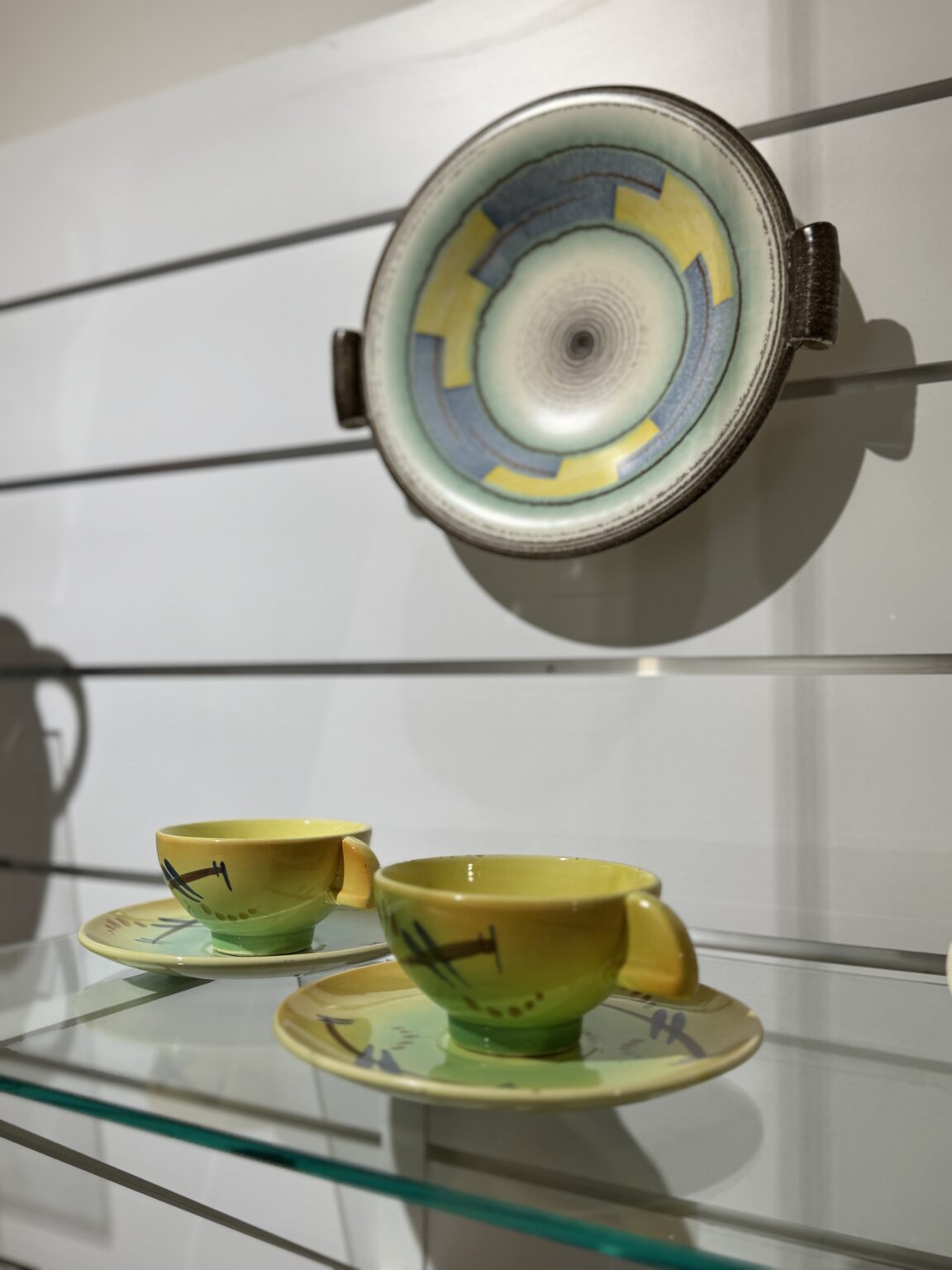 Dwie żółto-zielone filiżanki na spodeczkach, nad nimi wisi talerz z żółto-błękitnymi wzorami.