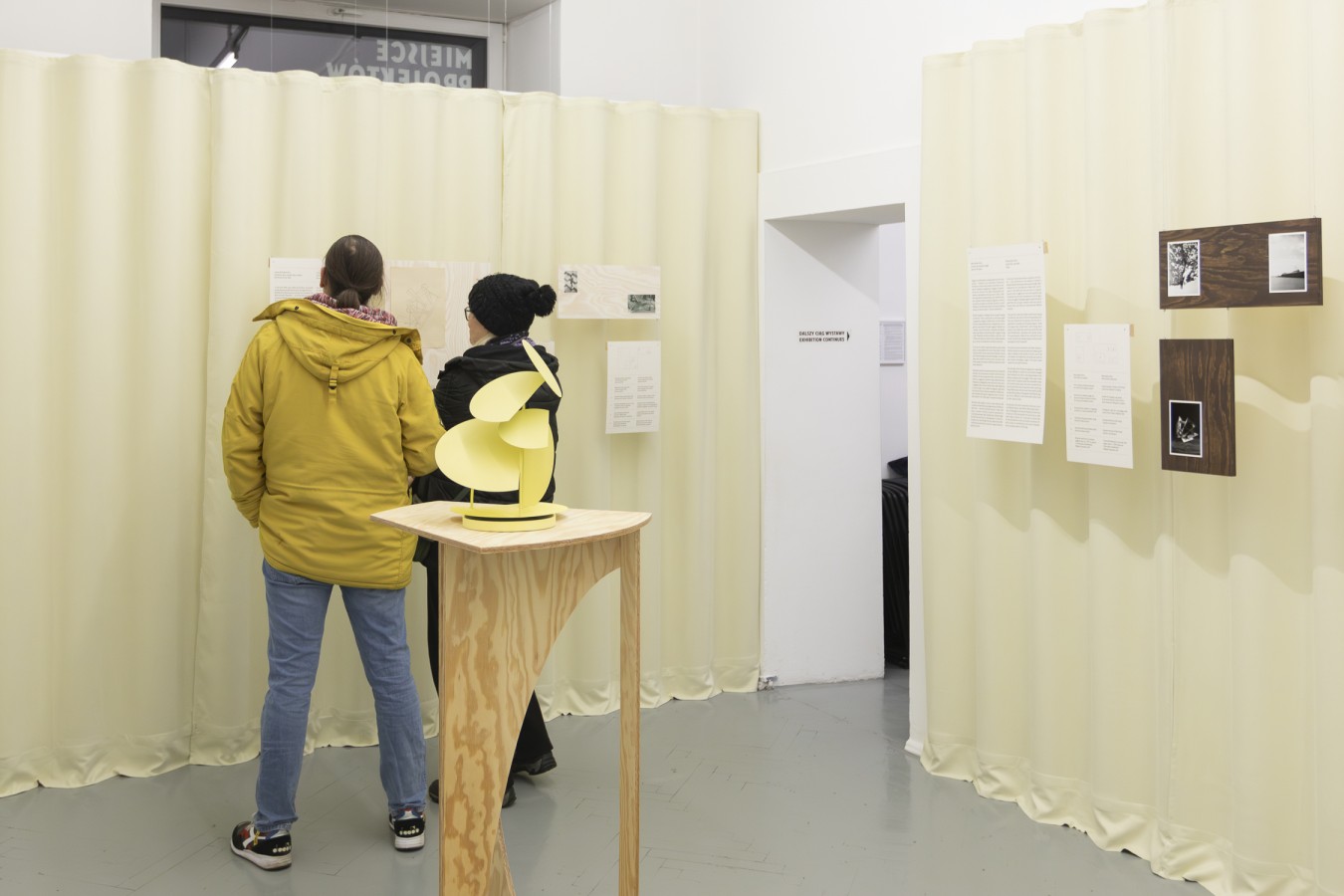 Wnętrze galerii. Na pierwszym planie postument z niewielką, żółtą, geometryczną rzeźbą. W tle kremowe kotary i stojąca tyłem para zwiedzających.
