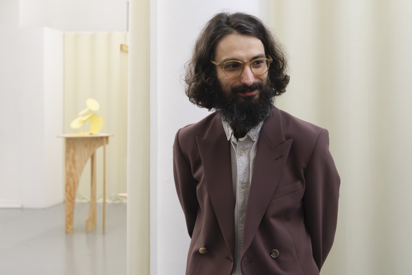 Na tle kremowych kotar uśmiechnięty mężczyzna o półdługich ciemnych, kręconych włosach i z gęstą ciemną brodą. Nosi okulary i brązową marynarkę.