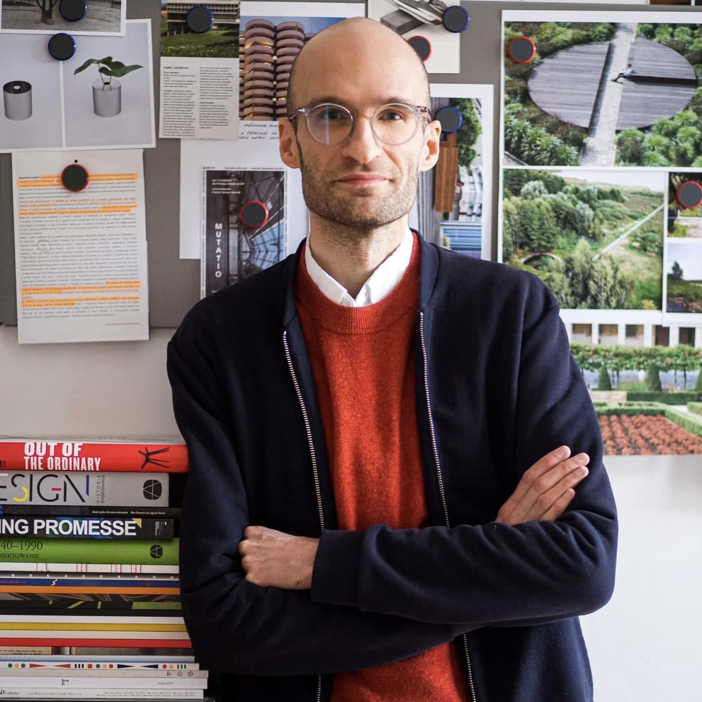Portret łysego mężczyzny w okularach, z rękami założonymi na piersiach. W tle na ścianie wizualizacje projektów i stos książek.