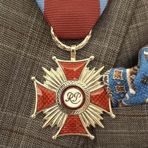 Zbliżenie na Krzyż Zasługi - czerwono-srebrny medal. Krzyż przypięty na przypięty na szarej marynarce w kratkę.