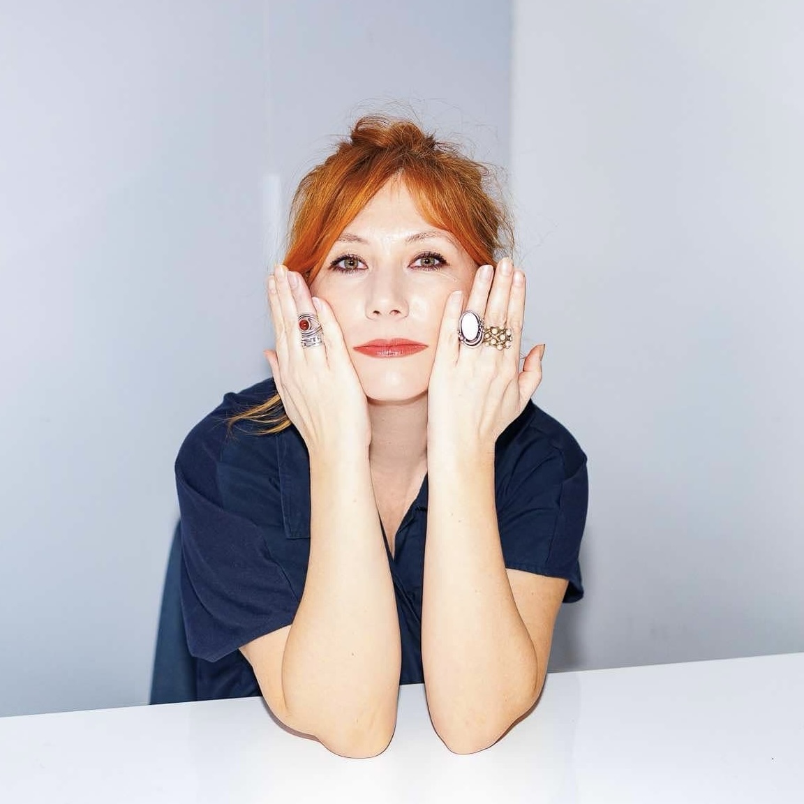 Portret rudowłosej kobiety na białym tle. Ubrana w granatową bluzkę, opiera łokcie o biały stół, przy twarzy trzyma dłonie ozdobione dużymi pierścieniami.