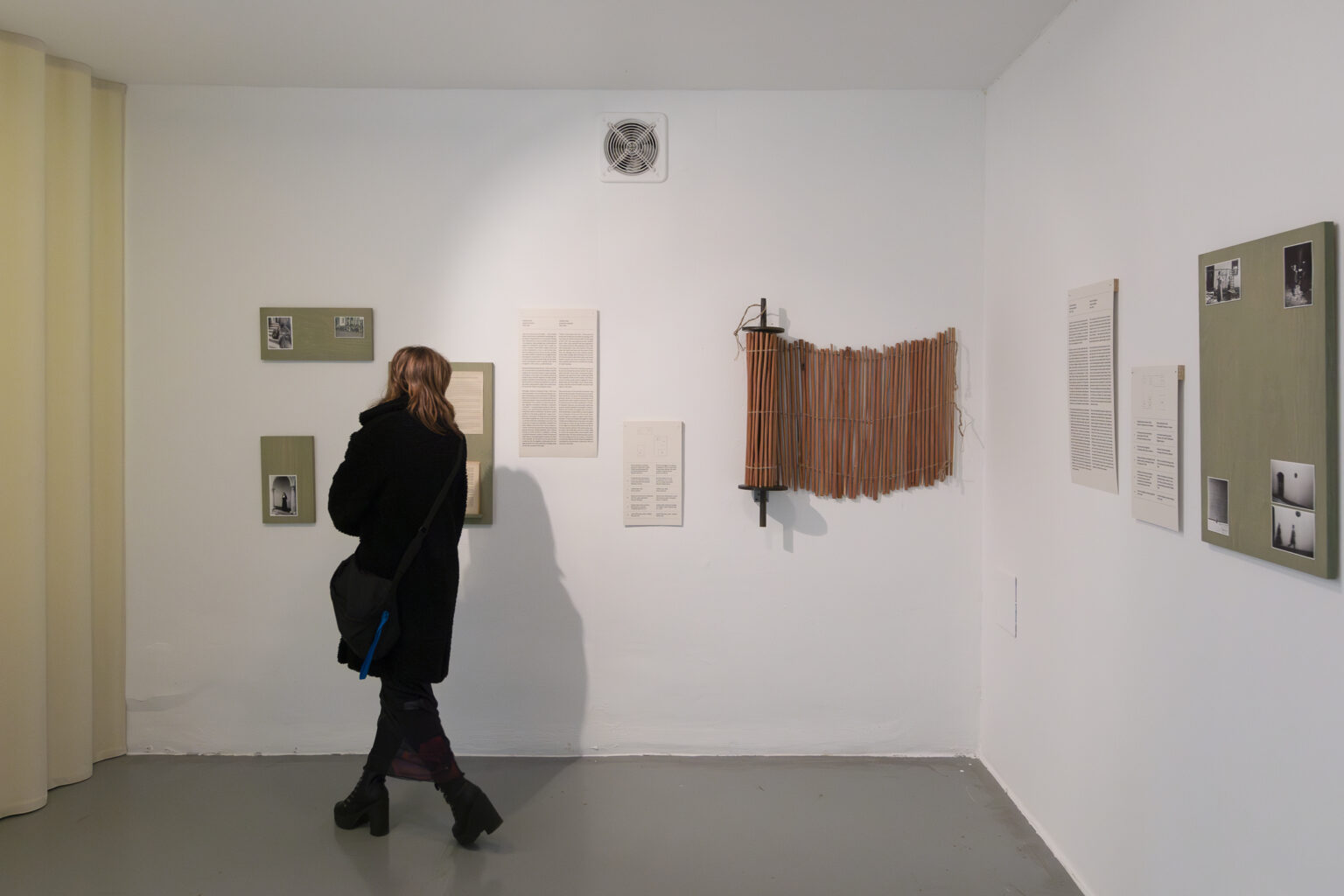 Wnętrze galerii. Na białej ścianie kilka fotografii, tekstów i obiekt przypominający nawinięte na wałek ogrodzenie z cienkich patyczków. Przed ścianą tyłem do obiektywu stoi kobieta.