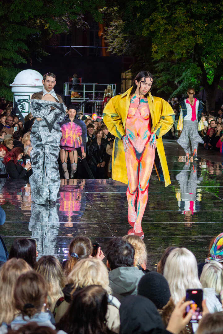 Scena po zmroku. Na wybiegu o lustrzanej powierzchni kilkoro modeli. Na pierwszym planie młody mężczyzna w obcisłym kombinezonie pomalowanym jak kobiece ciało, na to narzucony żółty płaszcz.